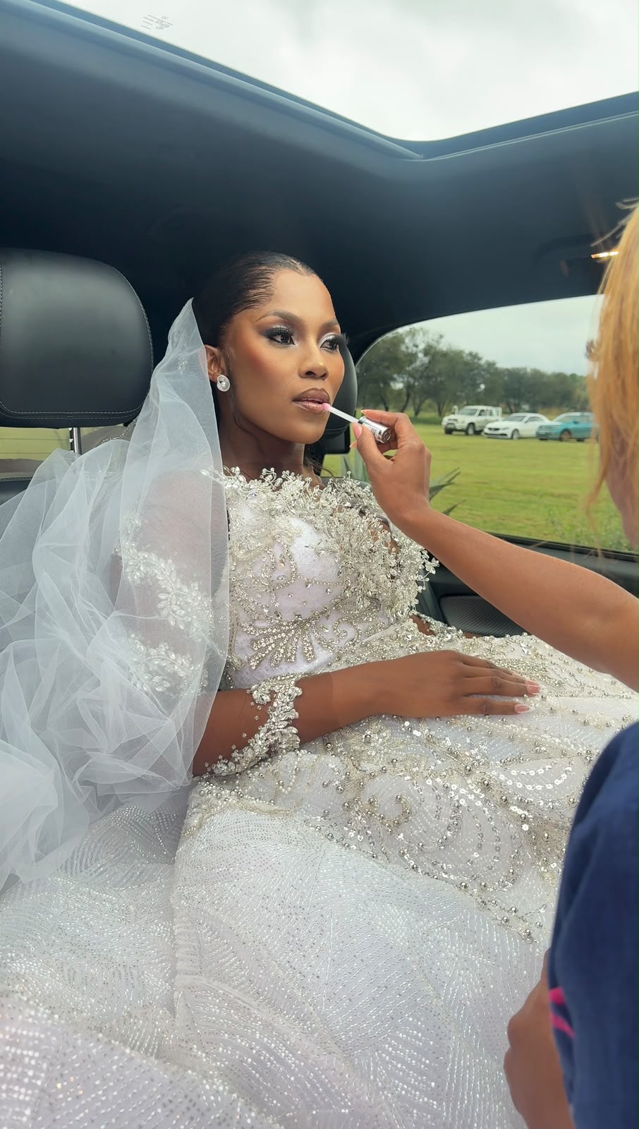 Bride in a beaded gown and veil having gloss applied to her lips, seated in a car on her wedding day